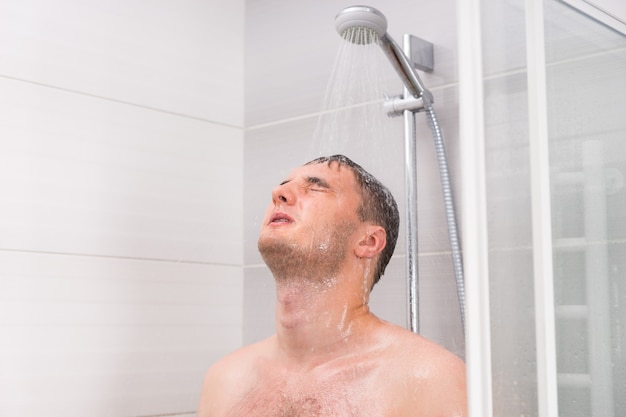 Young man with closed eyes taking a shower, standing under flowing water in shower cabin with transparent glass doors in the bathroom