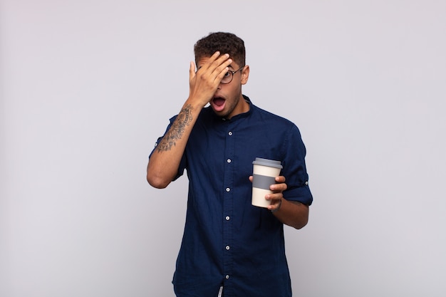 Young man with a coffee looking shocked, scared or terrified, covering face with hand