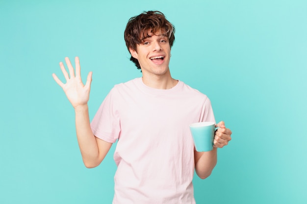 Young man with a coffee mug smiling happily, waving hand, welcoming and greeting you