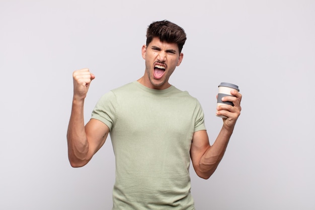 Young man with a coffee shouting aggressively with an angry expression or with fists clenched celebrating success