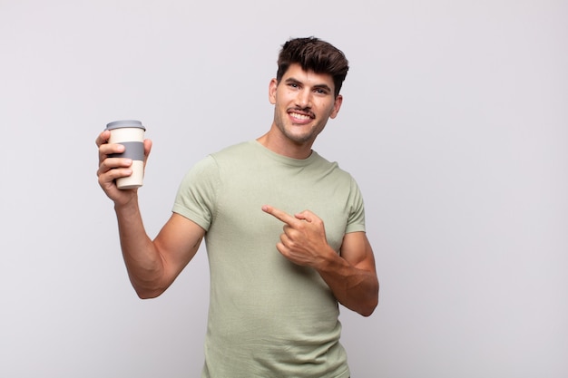 young man with a coffee smiling cheerfully, feeling happy and pointing to the side and upwards, showing object in copy space