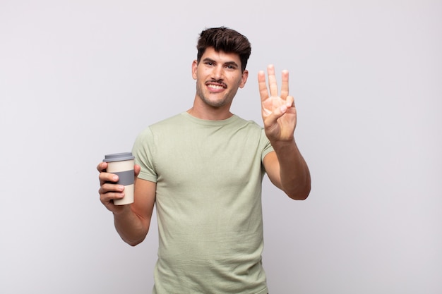 Young man with a coffee smiling and looking friendly, showing number three or third with hand forward, counting down