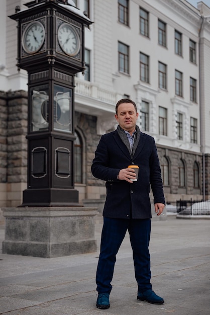 A young man with coffee stands in a city square with a clock tower waiting for a meeting fulllength vertical portrait