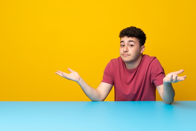 Young man with colorful wall and table having doubts while raising hands