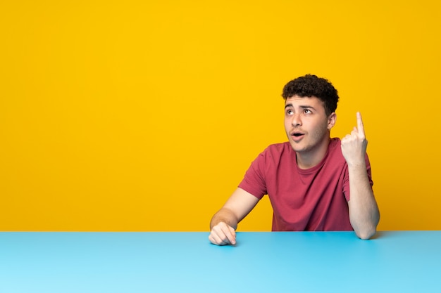 Young man with colorful wall and table pointing up and surprised