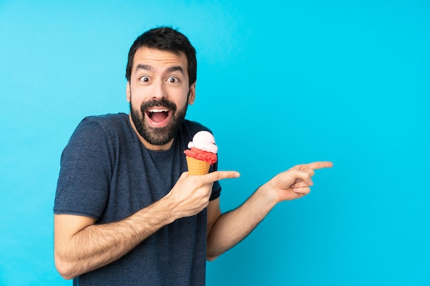 Young man with a cornet ice cream over isolated blue wall surprised and pointing side