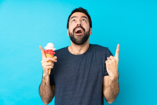 Young man with a cornet ice cream over isolated blue wall surprised and pointing up