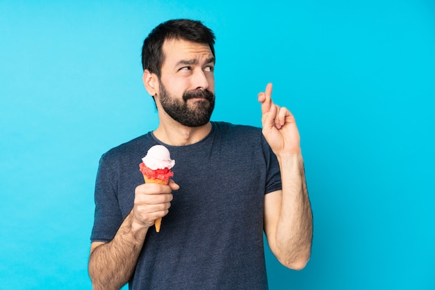 Young man with a cornet ice cream over isolated blue wall with fingers crossing and wishing the best