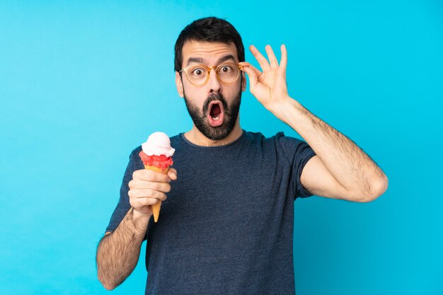 Young man with a cornet ice cream over isolated blue wall with glasses and surprised