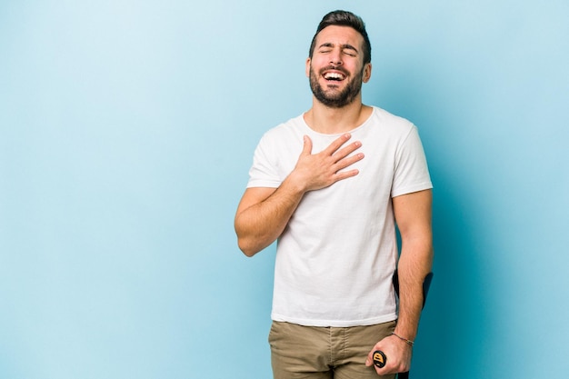 Young man with crutches isolated on blue background laughs out loudly keeping hand on chest