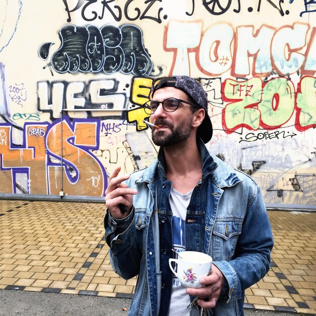 Photo young man with cup smoking cigarette against graffiti wall