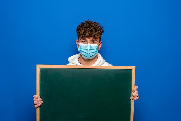 Young man with curly hair dressed in a white sweatshirt and surgical mask holding a blank white board isolated on blue background