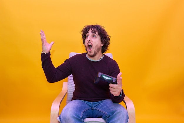 Young man with curly hair playing video games over isolated yellow background. Surprised and protesting looking to the game.