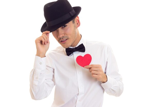 Young man with dark hair in white Tshirt and bowtie holding a red paper heart in his hands