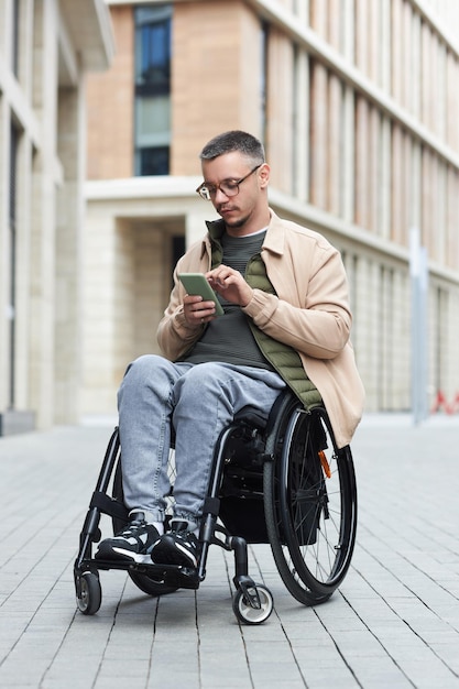 Молодой человек с инвалидностью сидит в инвалидной коляске и отправляет текстовое сообщение по телефону