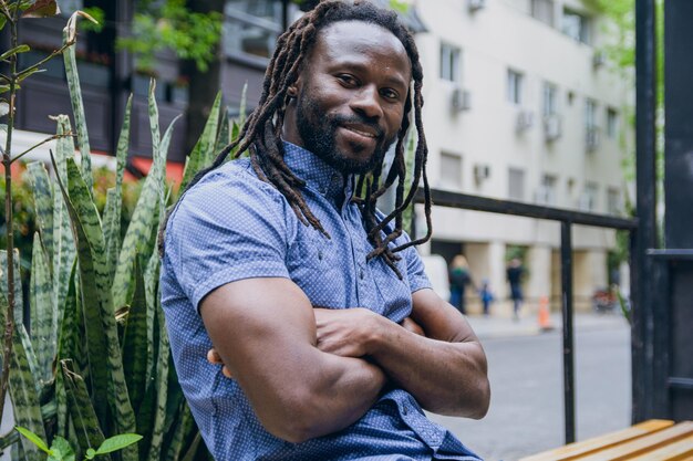 Photo young man with dreadlocks and beard in casual clothes sitting with arms crossed outside cafe happy and smiling looking at camera