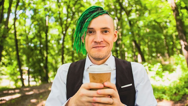 Young man with dreadlocks drinking coffee in forest park Stylish hipster in business suit with green hairstyle enjoying hot drink from paper cup in summertime