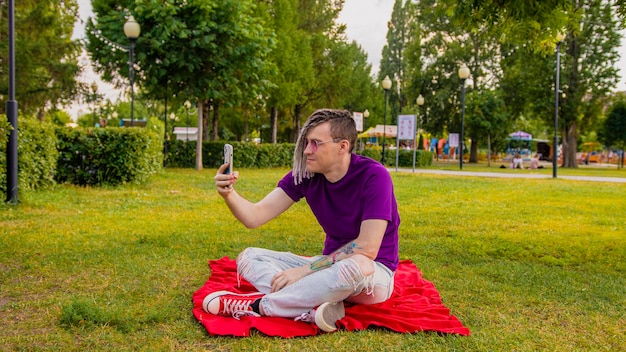 ドレッドヘアを持つ若い男が公園の毛布に座ってスマートフォンで自分撮りをしているハンサムな男が緑の芝生に座って晴れた日に自分の携帯電話を使って自分撮りをしている