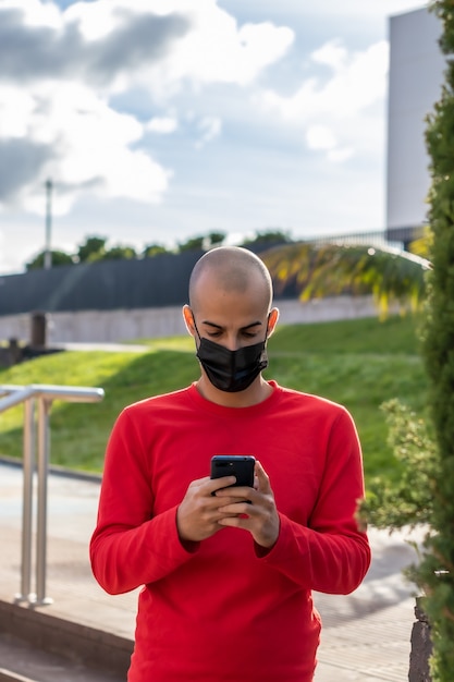Молодой человек в маске проверяет свой мобильный телефон и разговаривает в парке