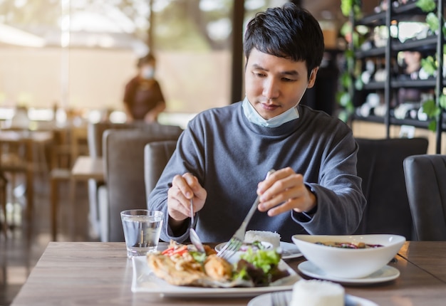 식당에서 음식을 먹는 얼굴 마스크를 가진 젊은 남자, 코로나 바이러스 (Covid-19) 대유행을 보호하기위한 새로운 일반 개념