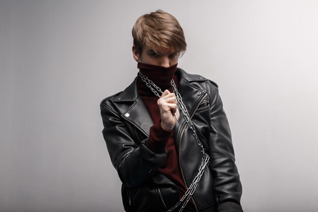 young man with a fashionable hairstyle in red golf in a black stylish jacket in black jeans holds a metal chain in his hands and looks into the camera against the background of a white wall. cute guy