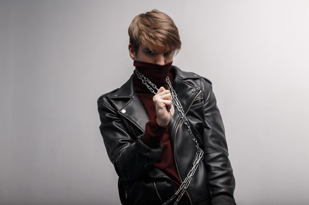 Young man with a fashionable hairstyle in red golf in a black stylish jacket in black jeans holds a metal chain in his hands and looks into the camera against the background of a white wall. Cute guy