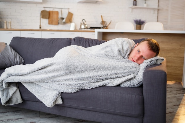 Young man with flu, sitting on the couch at home, wrapped in a blanket.