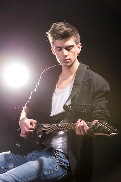 Young man with guitar in dark room with lights behind him.