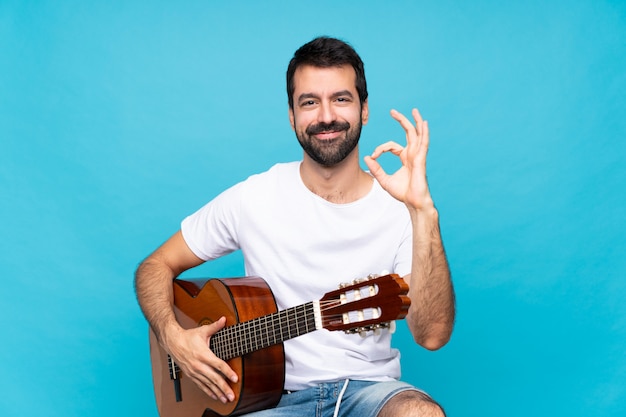 Young man with guitar over isolated blue showing ok sign with fingers