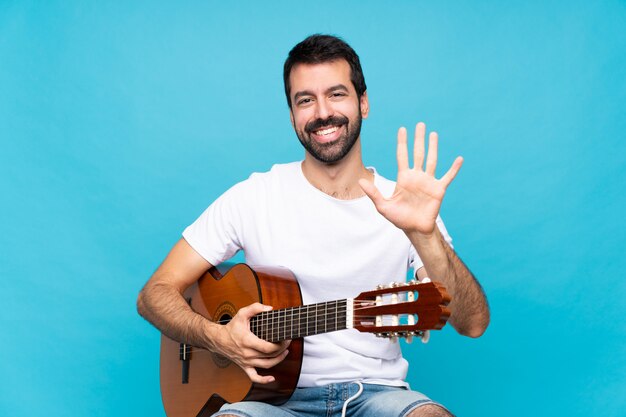 Young man with guitar over isolated blue wall counting five with fingers