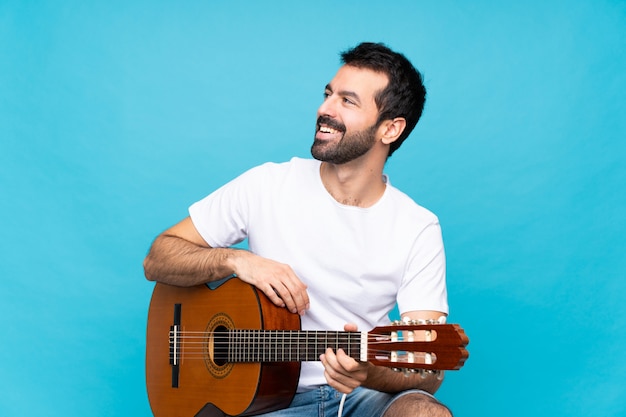 Young man with guitar over isolated blue wall happy and smiling