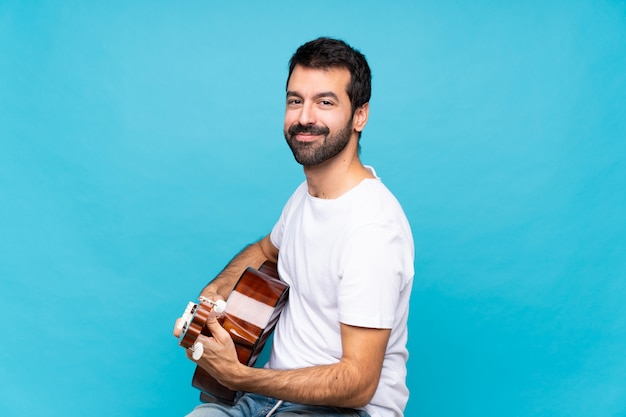 Young man with guitar over isolated blue wall with arms crossed and looking forward