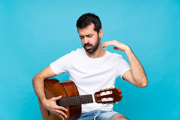 Young man with guitar over isolated blue with tired and sick expression