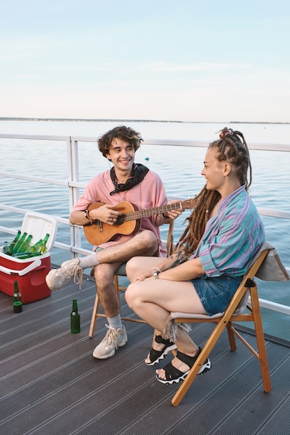 Young man with guitar looking at his girlfriend while both relaxing on pier