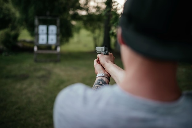 Young man with hat holding gun and shooting to target in countryside at daylight rear view