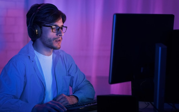 ヘッドホンとメガネをかけた若い男が紫と青で照らされた部屋でコンピューターに座っています