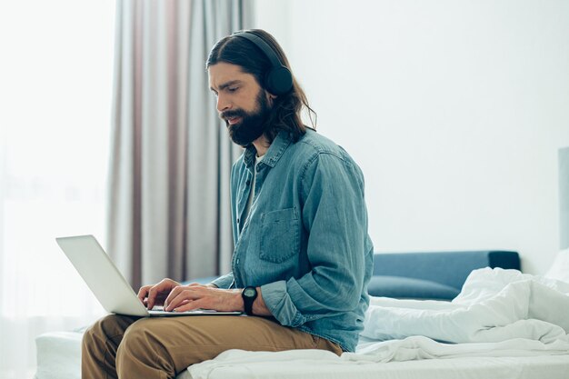 Young man with headphones on his head looking at the screen of a modern laptop while sitting on the bed with it