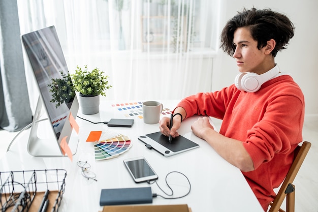 young man with headphones looking at computer screen while using graphics tablet and stylus to retouch photos