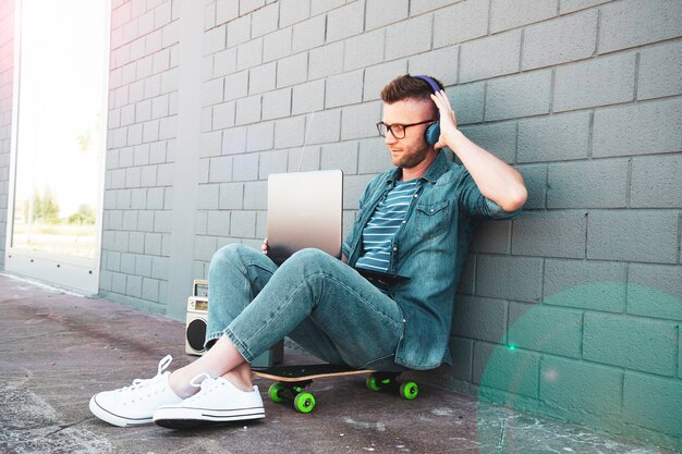 Young man with headphones using laptop in the street trendy\
cool guy having fun surfing online and listening music influencer\
or blogger working outdoors technology and fashion lifestyle\
concept