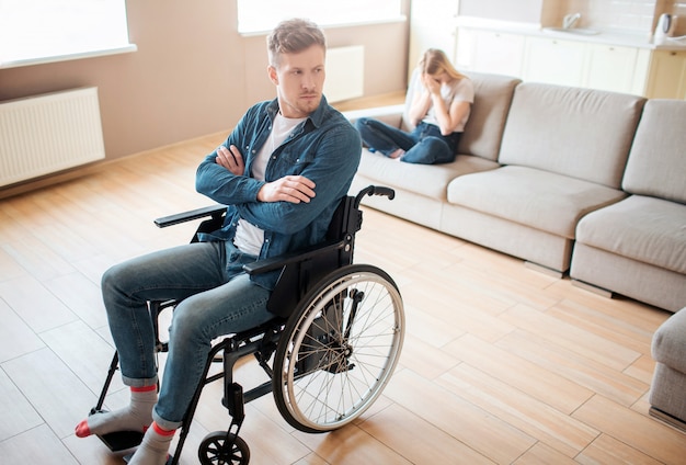 Young man with inclusiveness and disability sit on wheelchair in front. Hands crossed and upset. Young woman sit behind on sofa and cry. Emotional sickness and stress.