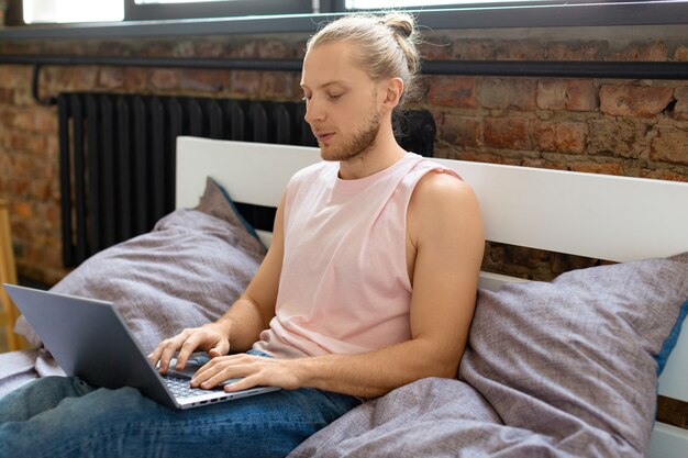 日常生活の中で現代の技術の概念のベッドでラップトップを持つ若い男