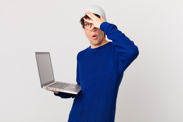 Young man with a laptop looking shocked, scared or terrified, covering face with hand