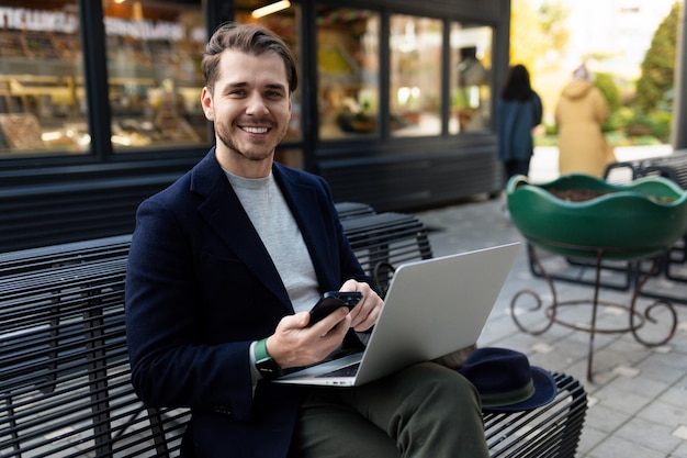 A young man with a laptop and a phone sits on a bench next to a city cafe on the street principles