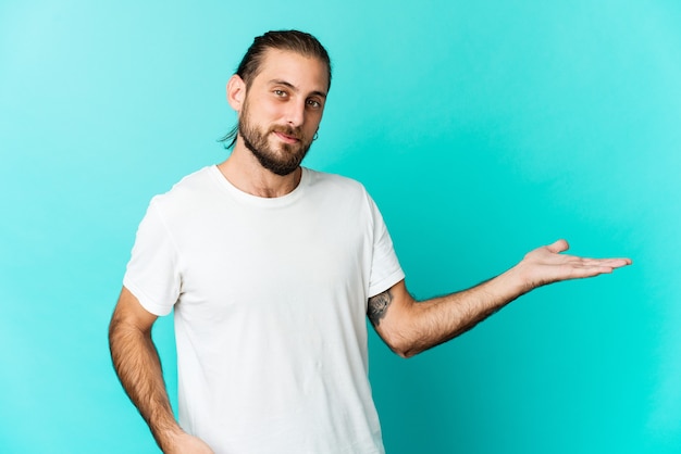 Young man with long hair look showing a copy space on a palm and holding another hand on waist.