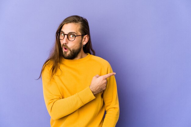 Young man with long hair look smiling and pointing aside, showing something at blank space.