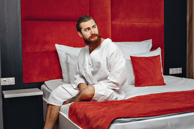 Young   man with a lush beard and long hair sitting in a light robe on a bed