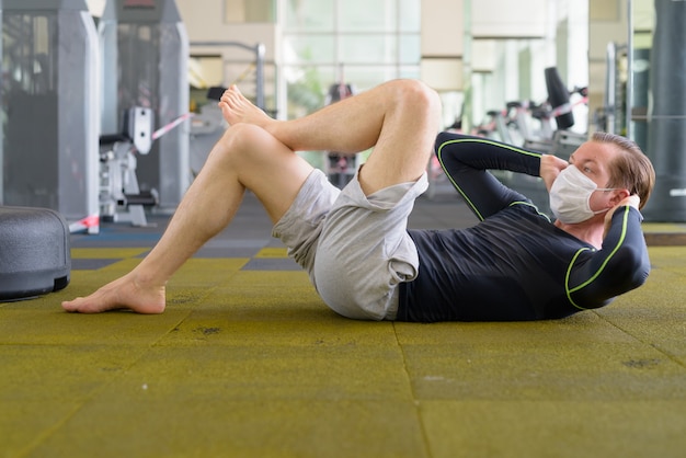 young man with mask doing sit ups on the floor at gym during coronavirus covid-19