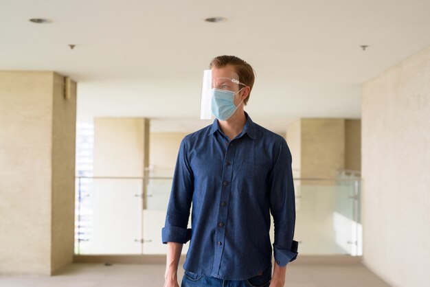 Young man with mask and face shield for protection from coronavirus outbreak thinking at modern building