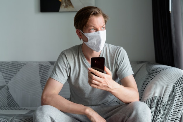 Young man with mask thinking while using phone at home under quarantine