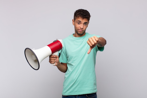 Young man with a megaphone feeling cross, angry, annoyed, disappointed or displeased, showing thumbs down with a serious look